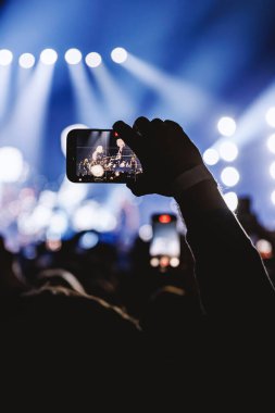 Parlak mavi sahne ışıkları altında canlı müzik çalan bir bandı yakalamak için bir izleyicinin elinde yakın plan bir fotoğraf..