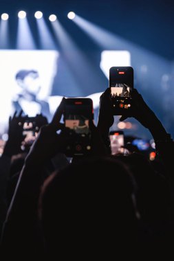 Birkaç izleyici, parlak spot ışıkları ve büyük bir video ekranı olan karanlık bir mekanda canlı performans kaydetmek için aynı anda akıllı telefonlarını kaldırıyor..