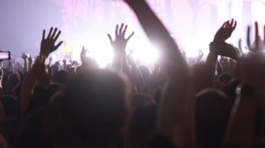 Canlı müzik konserinde büyük bir kalabalık heyecanla ellerini kaldırıyor. Sahnede parlak sahne aydınlatmaları, mercek parlamaları ve ışıldayan bir arka plan karşısında seyircilerin siluetleri yer alıyor..