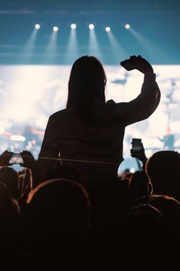 Siluetli bir insan daha iyi bir görüntü elde etmek ve sahnenin bir videosunu kaydetmek için kalabalığın üzerinde oturur. Arka plan serin mavi ve beyaz sahne ışıklandırması içeriyor.