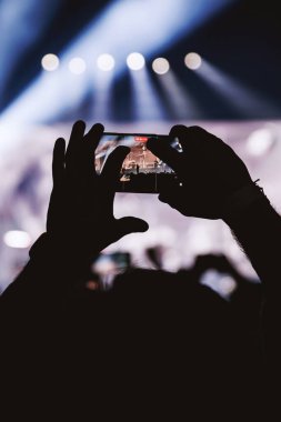 Telefonu yatay olarak tutan ellerin yakın çekimi, ekranda bandın net bir görüntüsünü gösteriyor. Sahne serin mavi ışıklar ve beyaz spot ışıklarıyla aydınlanır..