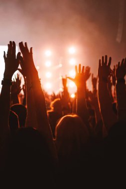 Hayranların siluetleri heyecan verici bir müzikal performans sırasında kollarını sahneye uzatırlar. Parlak, sıcak spot ışıkları arka planda yoğun bir şekilde parlıyor ve yüksek enerjili bir titreşim yaratıyor..