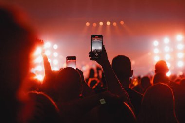 Kalabalıktaki bir kişinin silueti, parlak kırmızı spot ışıklarıyla dolu bir sahnede müzik performansı kaydetmek için cep telefonu kullanıyor..