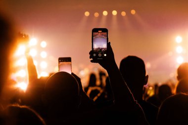 Kalabalığın çok üstünde tutulan bir akıllı telefon sahnenin geniş bir görüntüsünü yakalıyor. Sıcak altın ışık arenayı dolduruyor, enerjik bir yaz festivali havası yaratıyor..