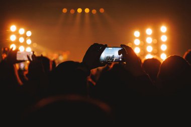 Bir müzik etkinliğinde, sahneyi yakalamak için çok sayıda akıllı telefonun tutulduğu geniş açılı bir çekim. Parlak, sıcak spot ışıkları canlı, yüksek enerjili bir atmosfer yaratır..