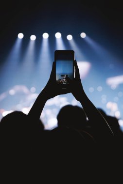 Bir hayran, akıllı telefon kullanarak canlı bir şovun fotoğrafını ya da videosunu çeker. Arka planda dramatik mavi spot ışıkları olan minimalist kompozisyon.