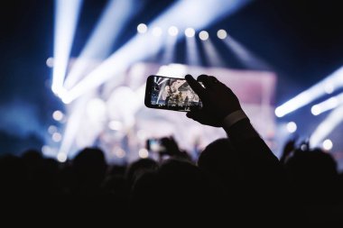 Kalabalığın yakın çekimi, sahnede canlı performans sergileyen bir grubu kaydetmek için elinde cep telefonu olan birini gösteriyor. Parlak mavi ışıklar, karanlık konser salonunda dramatik bir atmosfer yaratır..