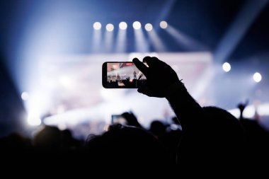 Seyirci, beyaz ve mavi ışıklarla dolu parlak bir sahnede canlı performans çekmek için akıllı telefonu yatay olarak kullanır..