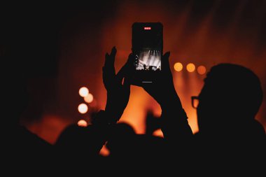 Kalabalıktan biri, sahnedeki sıcak turuncu ışıklandırmalı bir performansın fotoğrafını çekmek için telefonunu dikey olarak kaldırıyor..