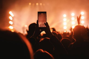 Canlı müzik şovu kaydetmek için bir hayranın tuttuğu akıllı telefon. Arka planda parlak turuncu spot ışıkları ve büyük bir kalabalığın siluetleriyle puslu sahne atmosferi yer alıyor..