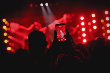 Kalabalıktaki bir kişinin silueti, canlı kırmızı ışıkların olduğu bir sahnede müzik programı kaydetmek için bir akıllı telefonu dikey olarak tutuyor..