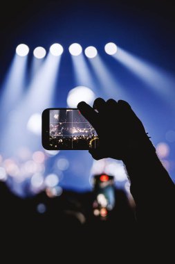 Canlı yayını kaydetmek için cep telefonu tutan bir elin siluetini gösteren dikey bir çekim. Serin mavi spot ışıkları sahneyi yukarıdan aydınlatıyor..