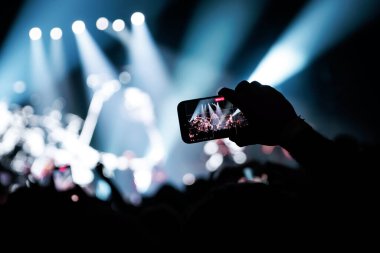 Canlı yayın yapan bir akıllı telefon ekranının yatay görüntüsü. Mekan, modern hayranların dijital kayıt deneyimlerini vurgulayan havalı mavi spot ışıklarıyla aydınlanıyor..