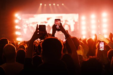 Birkaç kişinin bir grubu kaydetmek için cep telefonlarını tuttuğu bir seyircinin arka görüntüsü. Sahne, sahne ışıklarından gelen sıcak turuncu ve kırmızı bir parıltıyla dolu..