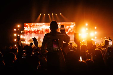 Merkezi bir şahsiyet siluetli kalabalığın ortasında oturmuş canlı yayını kaydetmek için telefonları tutuyor. Sıcak turuncu ışık çarpıcı bir yüksek kontrastlı zemin oluşturur.