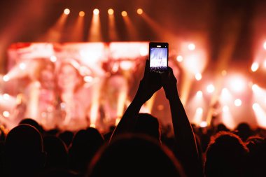 Parlak turuncu sahne ışıklandırması olan bir arenada müzik performansı çekmek için telefonu dikey tutan kişinin merkezi bir silueti..