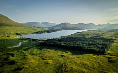 Yüksek açılı bir perspektif dalgalı yeşil tepeleri, yoğun çam ormanlarını ve berrak bir yaz gökyüzünün altında sakin bir gölü gösterir..