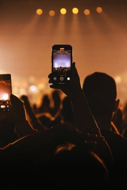 Canlı müzik performansı yakalayan parlak bir akıllı telefon ekranının hayran perspektifi. Görüntüde siluetli bir kalabalık, turuncu sahne ışıkları ve arka planda bokeh efektleri yer alıyor..