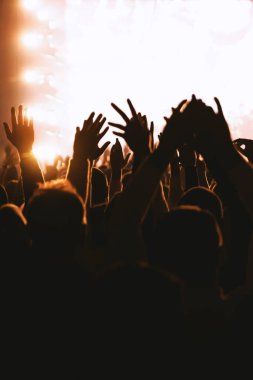 Konser izleyicileri tarafından derinden çekilen bir bakış açısı. Hayranların siluetleri, dinamik bir arkaplan yaratmak için canlı, parıldayan bir sahneye doğru ellerini kaldırır..