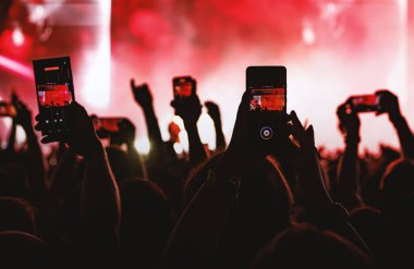 Konser hayranları kırmızı sahne aydınlatmalı bir gösteri sırasında bir performans kaydetmek için mobil cihazları kaldırıyor.