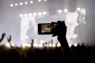 Siluetli bir insan, parlak bir sahnede çalan bandın videosunu çekmek için akıllı telefonu yatay olarak tutuyor..