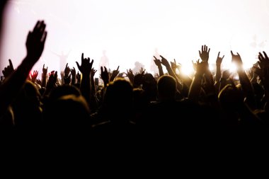 Siluetli hayranların yoğun beyaz sahne aydınlatması altında canlı bir konserde ellerini kaldırması geniş bir manzara..