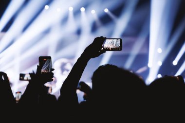 Mavi ve beyaz ışık huzmelerinin yer aldığı bir konseri kaydetmek için cep telefonunu yatay olarak tutan silüetli bir el..