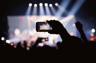 Canlı müzik performansını çekmek için bir el cep telefonunu tutuyor. Arka plandaki sahne birden fazla parlak spot ışığı ve ışınla aydınlatılıyor..