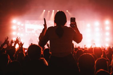 Canlı kırmızı ışıklar altında canlı performans kaydetmek için akıllı telefon kullanan bir kişinin silüeti..