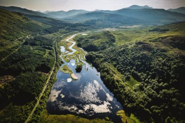 Geniş bir dron görüntüsü, orman tepeleri ve dağlarla çevrili yemyeşil bir vadiden geçen bir nehri yakalar. Su parlak gökyüzünü yansıtır, dingin ve doğal bir manzara yaratır..