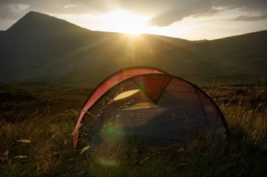 İskoçya 'da canlı bir günbatımı sırasında çimenli bir arazide küçük kırmızı bir çadır kurulur. Midges akşam ışığını yakalarken güneş uzak dağların tepelerinde parlıyor..
