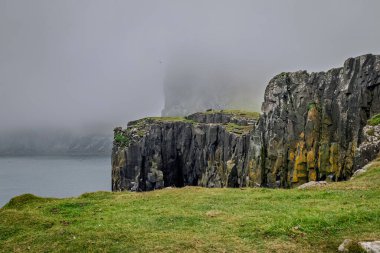 Dikey kaya oluşumları ve sivri kayalıklar yoğun bir sis örtüsünün altında okyanusla buluşuyor. Yeşil çimenler engebeli Highland kıyı şeridinin önünü kaplıyor..