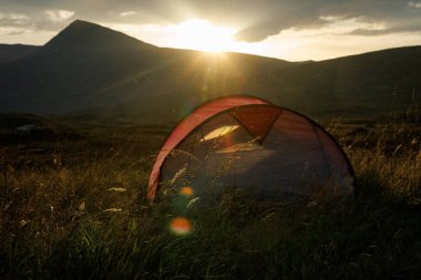 Bir insan dağlık arazide kamp yapıyor. Güneş bir dağ zirvesinin arkasında batıyor. Çimlerin üzerine altın bir işaret fişeği atıyor..