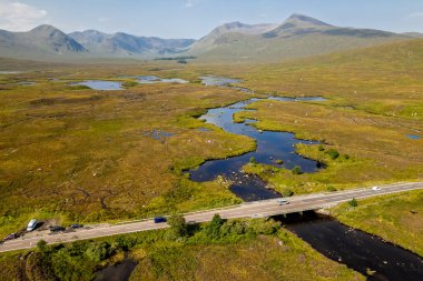 Yüksek açılı bir manzara, dağların arasındaki yeşil bir vadide arabaların geçtiği dolambaçlı bir nehir ve düz bir yolu gösteriyor..