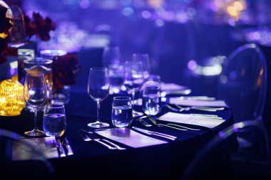 Kristal camdan yapılmış resmi yemek masası, cilalanmış çatal bıçak takımı ve atmosferik mavi sahne ışıkları altında menüler..