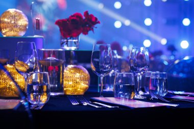 Bir ziyafet masasında kristal cam eşyalar, vazoda kırmızı güller ve altın küre lambaları bulunur. Arka plan mavi sahne ışıklandırması ve büyük bir mekânın bokeh efektleriyle dolu..