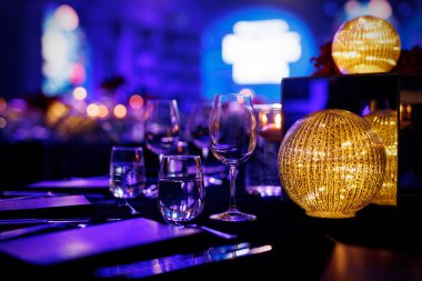 Bir gala yemeği için sehpada bardaklar ve cilalanmış çatal bıçaklar var. Ön planda büyük yuvarlak dokulu bir lamba parlak bir ekran ile bulanık mavi bir arka planda parlar..