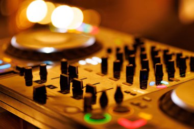 Çeşitli düğmeleri ve kaydırıcıları gösteren DJ mikser destesinin detaylı bir görüntüsü. Arka planda olay atmosferinden gelen yumuşak altın ışık çemberleri yer alıyor.