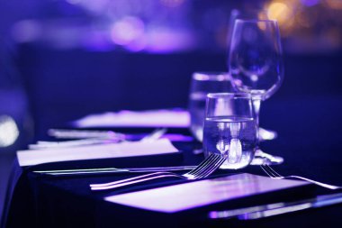 Yemek masasında mavi ve mor ışık yansımaları cam eşyalar ve cilalanmış çatal bıçaklar var..