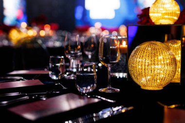 Altın ışıklı küresel bir lamba, parlatılmış cam eşyalar ve akşam bulanık bir arka plana karşı gümüş çatal bıçak içeren resmi bir gala masasının yakın çekimi..