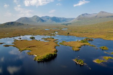 Geniş, mavi bir göl, küçük otlak adaları ve dağları gösteren geniş bir drone görüntüsü..
