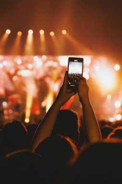 Kalabalığın dikey çekimi turuncu ışıkları yakalamak için akıllı telefon tutan bir silueti gösteriyor..
