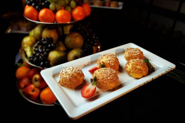 Beyaz dikdörtgen tabakta yuvarlak profiteroller var. Şekerle tozlanmış ve dilimlenmiş çilekle servis ediliyor..