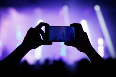 Parlak mor ışıklar ve kalabalığın altında sahnedeki bir müzik performansını çekmek için cep telefonu tutan bir el silueti..