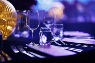 Bir kristal şarap kadehi ve bir su bardağı mavi ve mor ortam ışıkları altında koyu bir masa örtüsünün üzerinde otururlar. Yumuşak odak noktasında parlatılmış gümüş takımlar ve dekoratif bir altın lamba görülebilir..