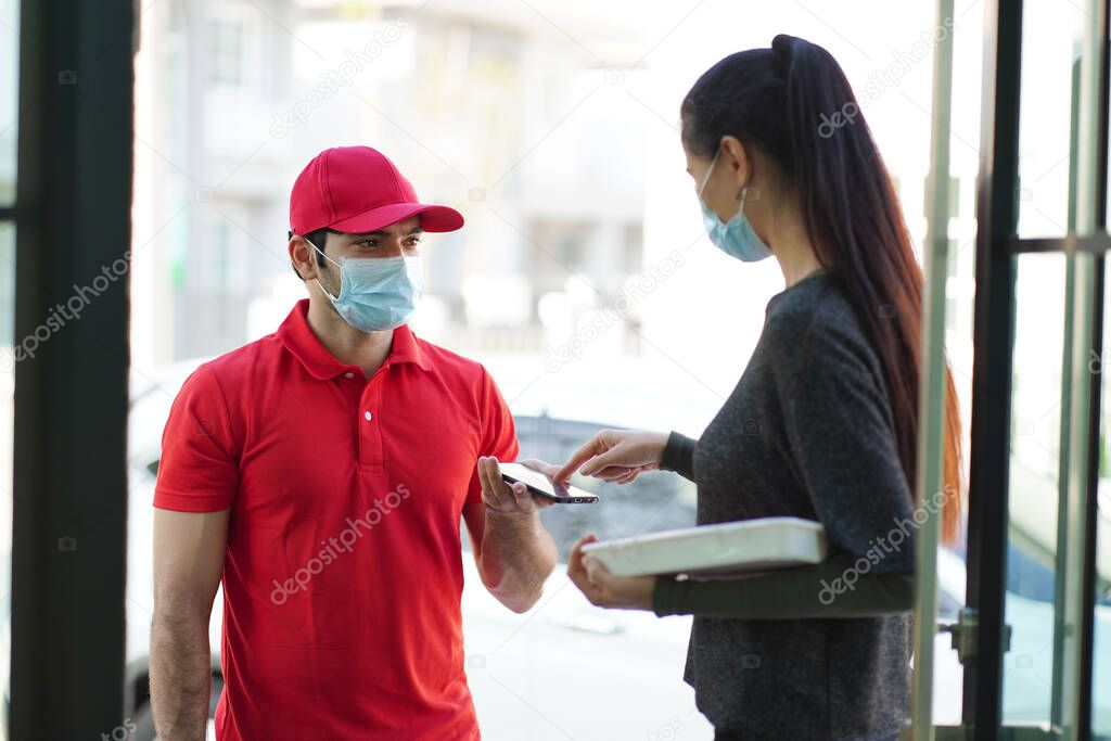 Beautiful customer wearing a surgical mask receives a food box from a food delivery man, wears a red uniform, and wears a mask delivered to the apartment. New normal concept. 
