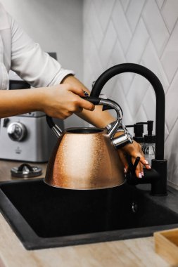 Bir kadın sıcak içecek hazırlamak için çaydanlıkta su toplar. Mutfağın içinde İskandinav tarzı