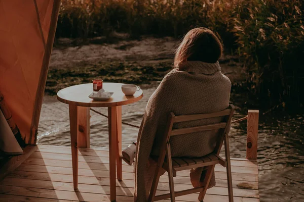 beautiful young girl drinks tea in nature by the lake. a woman is resting in a glamping in a picturesque location far from the city. self-isolation vacation without people