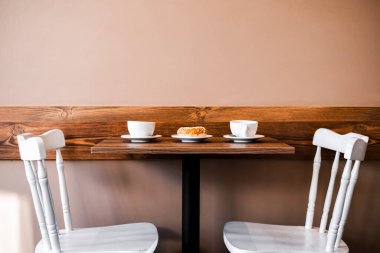 Kahve ve tatlı ahşap bir masada. Masanın arka planında iki beyaz sandalye var. Kahvaltı için rahat bir yer.