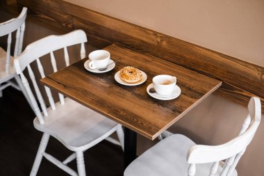 Kahve ve tatlı ahşap bir masada. Masanın arka planında iki beyaz sandalye var. Kahvaltı için rahat bir yer.